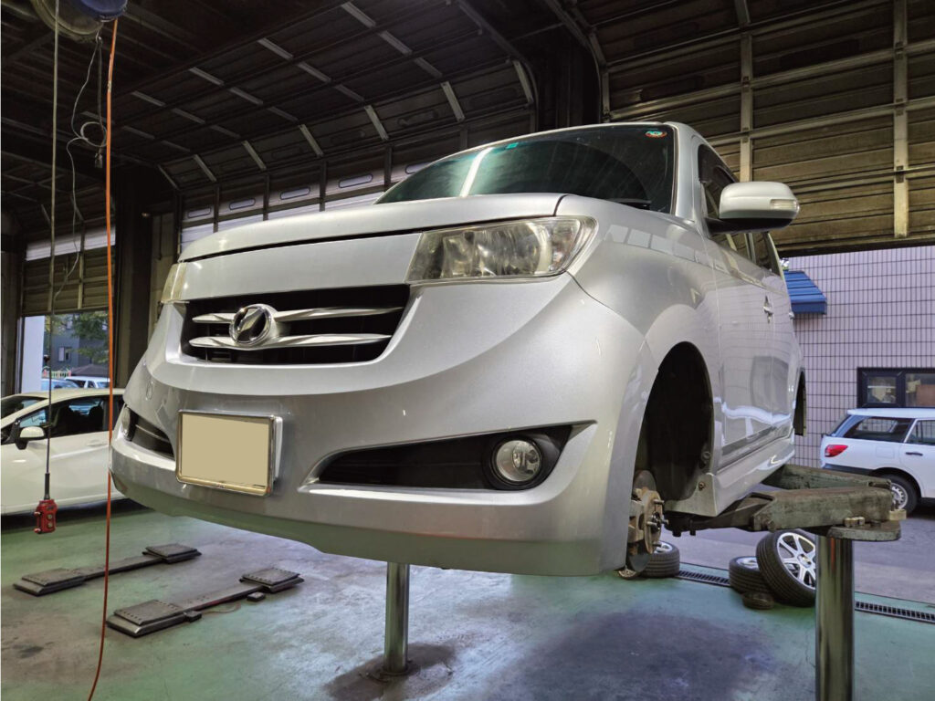 整備工場でリフトアップしタイやが外されているシルバーのトヨタBb